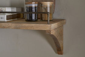 A close-up of the nkuku Meera Mango Wood & Iron Gallery Shelf - Natural, featuring a curved bracket and metal rail, holds glass jars with wooden lids filled with spices or tea leaves and books, set against a beige wall.