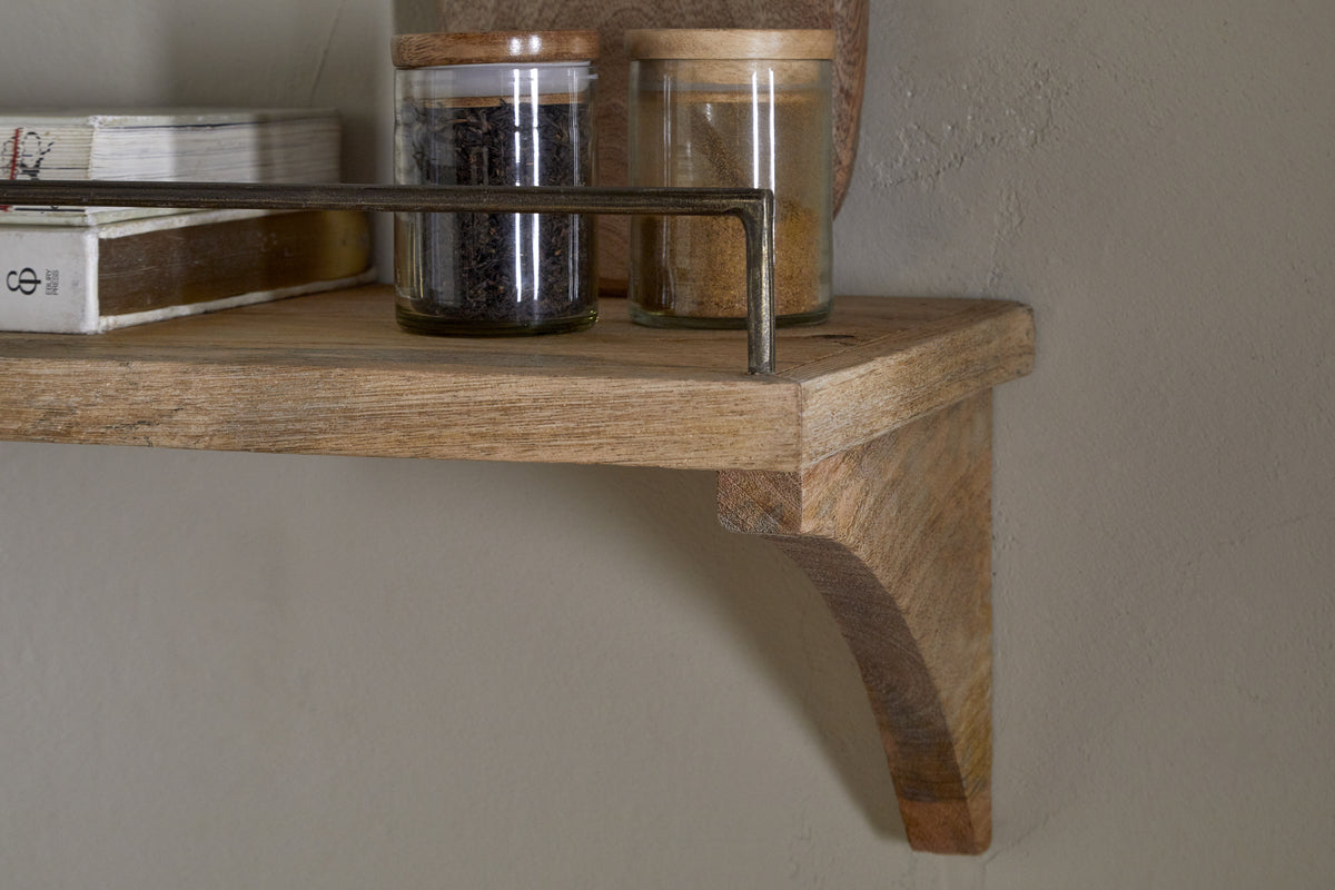 A close-up of the nkuku Meera Mango Wood & Iron Gallery Shelf - Natural, featuring a curved bracket and metal rail, holds glass jars with wooden lids filled with spices or tea leaves and books, set against a beige wall.