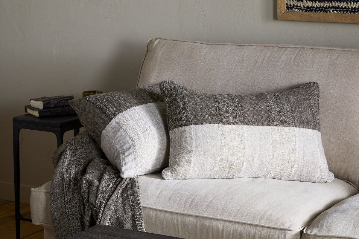 A beige couch with two nkuku Munir Linen Cushion Covers in charcoal—featuring a striped slubby texture—sits beside a black side table with stacked books, and a textured gray-and-white throw blanket draped over the arm.
