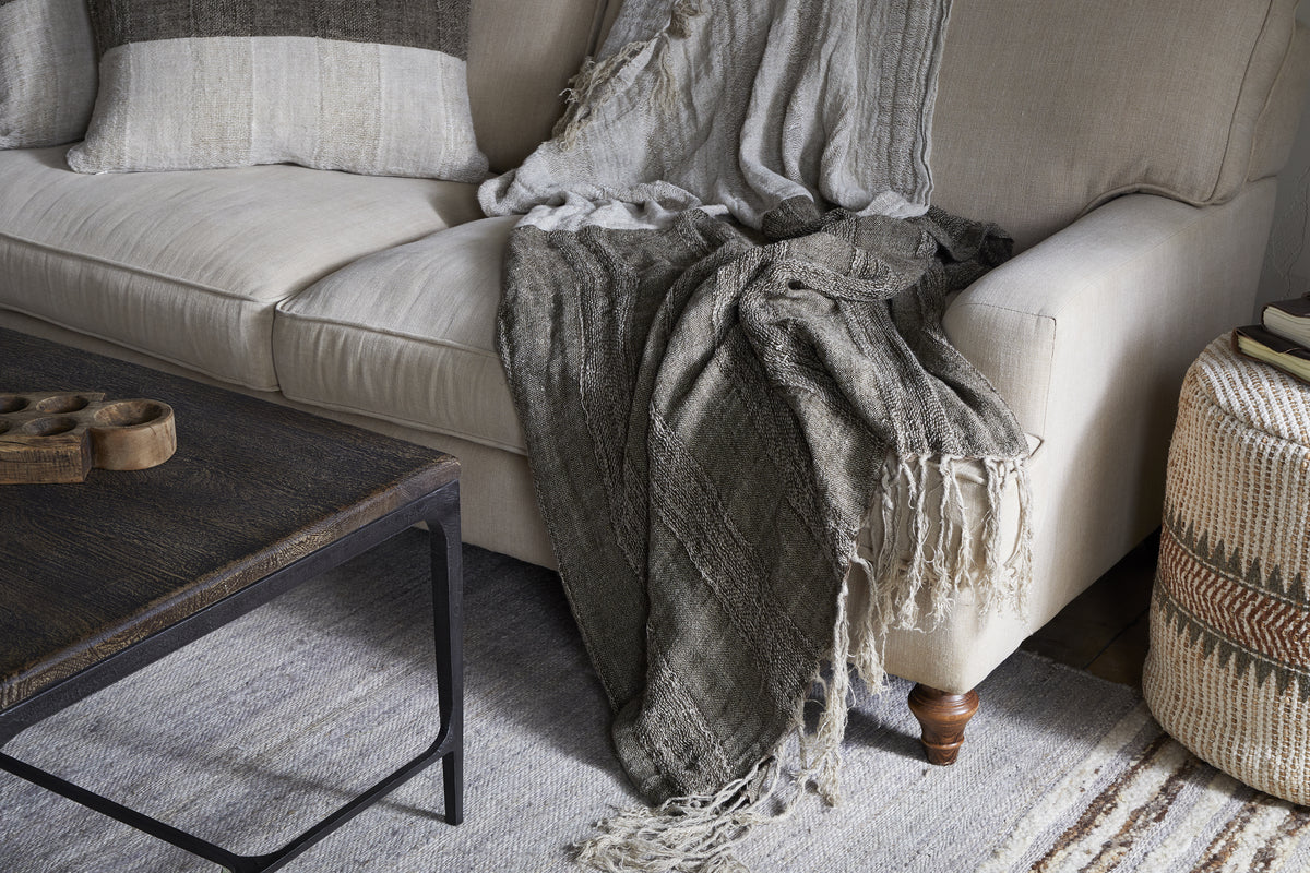A neutral-toned living room features a beige sofa draped with the nkuku Munir Linen Throw in Charcoal, textured gray and taupe throws, a matching pillow, dark wood coffee table, woven zigzag ottoman, and a light area rug on the floor.