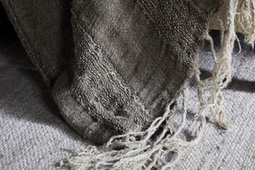 A close-up of the nkuku Munir Linen Throw in Charcoal draped over a gray rug, showcasing its slubby texture, frayed edges, and visible weave for a rustic, handmade feel.