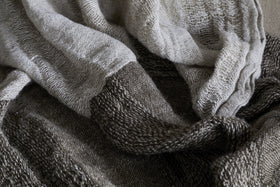 A close-up of the Munir Linen Throw in Charcoal by nkuku shows soft, textured wavy folds and wide alternating stripes in light beige and dark brown, highlighting its intricate knit pattern and cozy appeal.