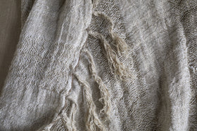 A close-up of the Munir Linen Throw - Charcoal by nkuku highlights its woven charcoal fabric with slubby texture. Frayed threads and decorative weave reveal organic, natural fibers and charming irregularities under the light.
