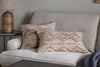 A beige sofa features two nkuku Nila Esha Cushion Covers in rust and indigo geometric patterns. A round black side table holds a brown ribbed lamp with a beige shade, while a blue knitted blanket is draped on the sofa’s armrest.