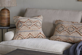 A beige sofa with two patterned throw pillows—one featuring the nkuku Nila Esha Cushion Cover in Rust & Indigo—sits by a round black side table topped with a ribbed brown lamp. A dark blue knit blanket is draped over the arm against a light gray wall.