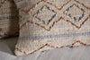 Close-up of the hand-embroidered nkuku Nila Esha Cushion Cover – Rust & Indigo, featuring textured diamond and line patterns on a beige base. It sits on a light beige surface with another similar cushion nearby.