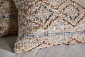 Close-up of the hand-embroidered nkuku Nila Esha Cushion Cover – Rust & Indigo, featuring textured diamond and line patterns on a beige base. It sits on a light beige surface with another similar cushion nearby.