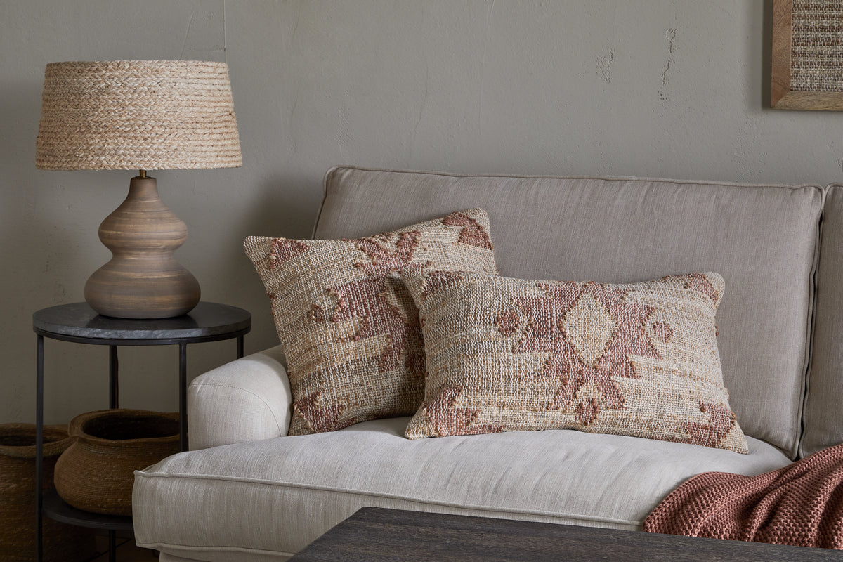 A beige couch features the nkuku Nila Gul Cushion Cover in Rust, paired with earthy-tone patterned pillows. A round black side table displays a woven lamp, baskets are below, and a rust knit blanket rests on the armrest.