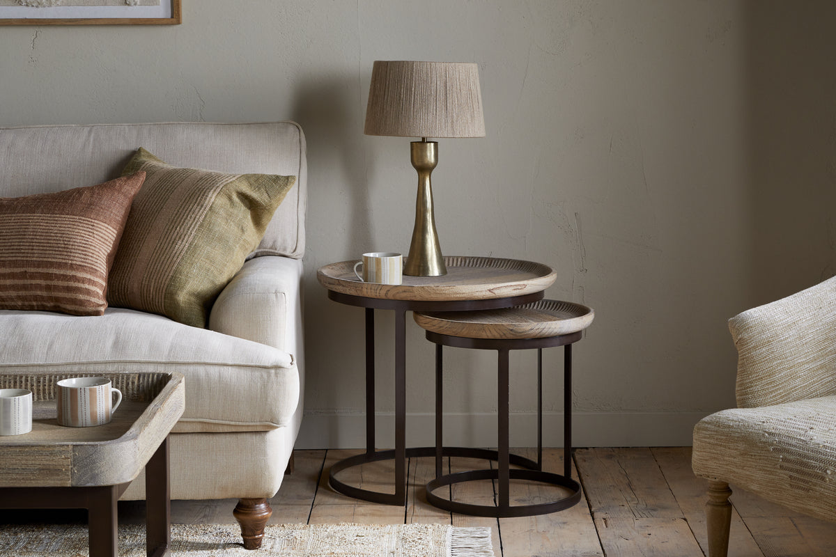 A cozy living room with a beige sofa, brown and green pillows, a gold lamp with beige shade, mugs, a small white candle, wooden floors, light walls, and nkuku’s Nisha Carved Reclaimed Wood & Iron Nesting Side Tables in Natural.