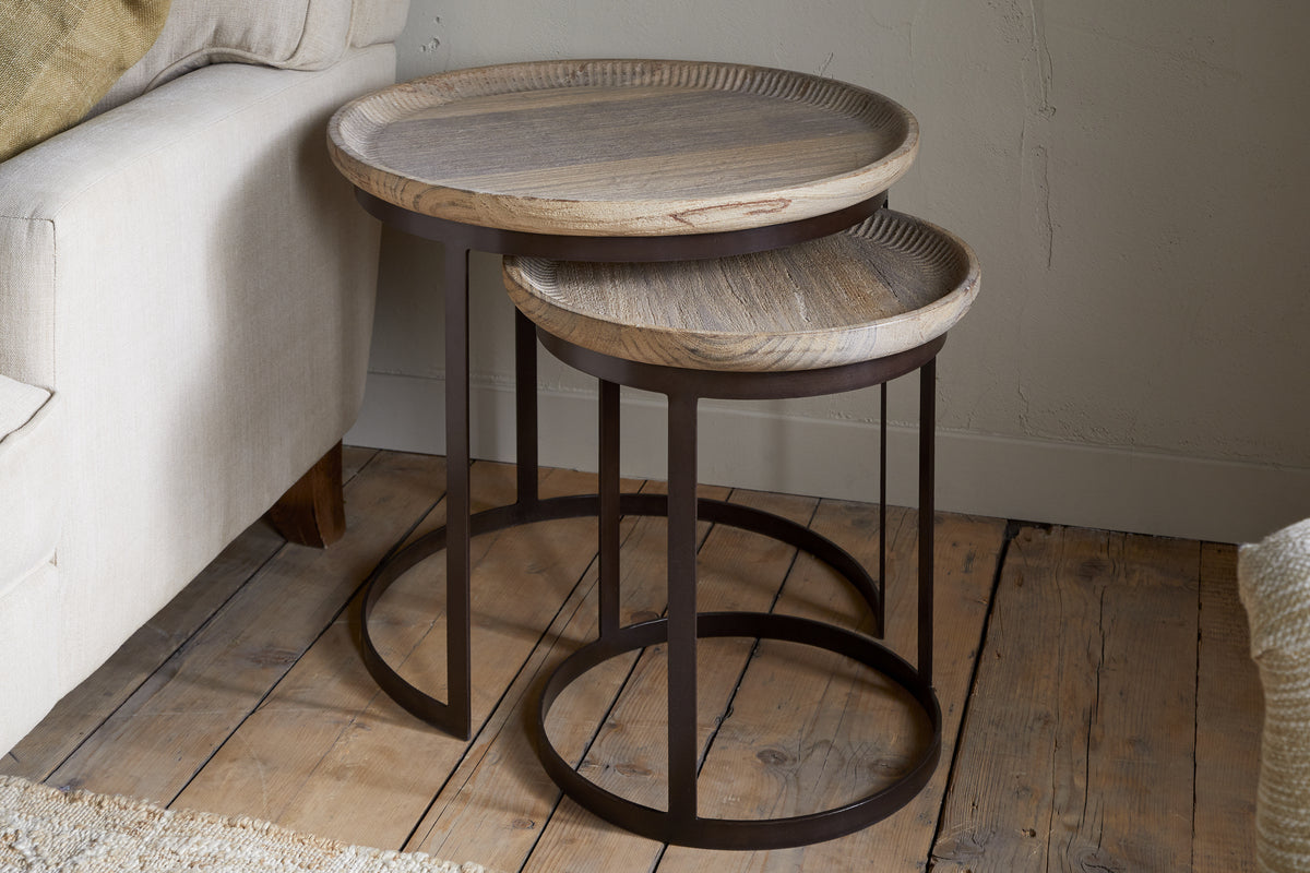 The nkuku Nisha Carved Reclaimed Wood & Iron Nesting Side Tables – Natural feature light wood tops and dark metal bases. The smaller table nests under the larger, set on a rustic wooden floor by a beige sofa and a textured light wall.