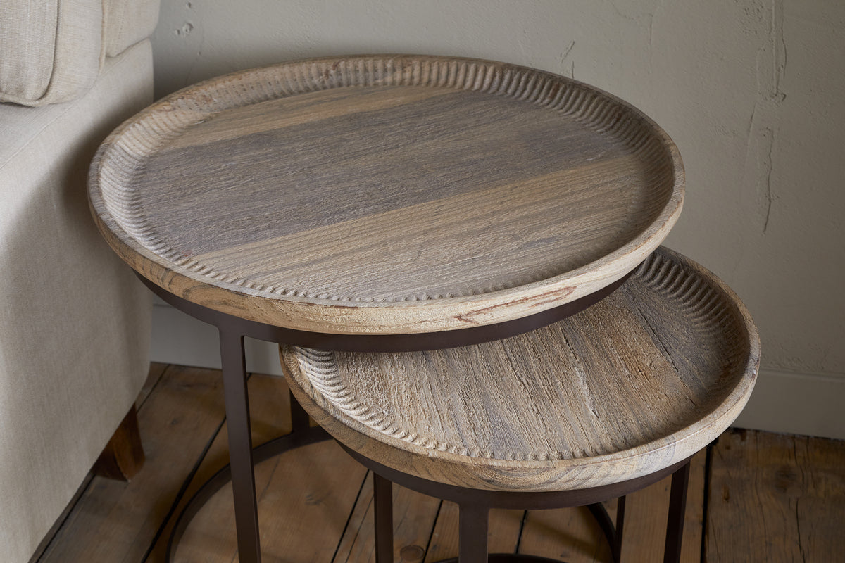 The Nisha Carved Reclaimed Wood & Iron Nesting Side Tables by nkuku, featuring a natural wood grain finish and metal legs, are arranged on a wooden floor beside a beige sofa with the smaller table partially tucked under the larger.