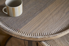 A close-up of the nkuku Nisha Carved Reclaimed Wood & Iron Nesting Side Table in Natural shows its textured light brown grain and decorative carved edge. A white mug with a vertical yellow and gray dotted line pattern sits near the upper left corner.