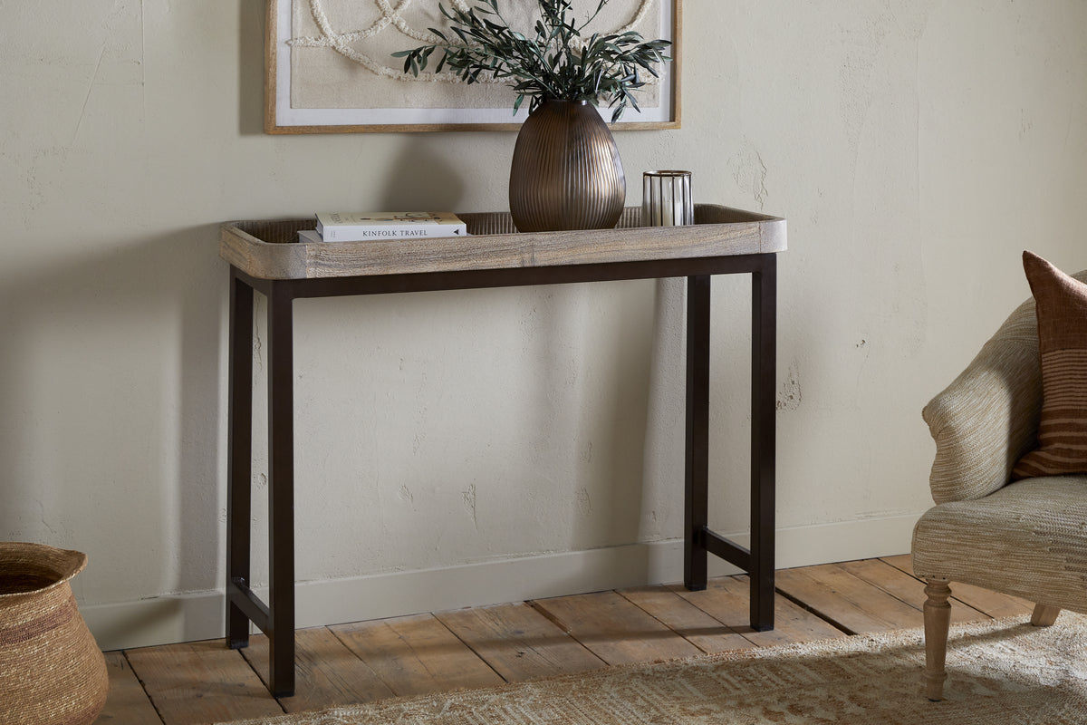 The nkuku Nisha Reclaimed Wood & Iron Console Table - Natural stands against a beige wall, topped with a brown vase of green branches, a glass candle holder, and a stacked white book. A beige cushioned chair is partially seen to the right.