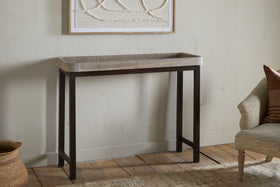 The nkuku Nisha Reclaimed Wood & Iron Console Table - Natural, featuring a tray top and dark iron legs, stands against a textured wall with an abstract framed artwork above and a woven basket plus beige upholstered chair nearby.
