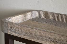 Close-up of the nkuku Nisha Reclaimed Wood & Iron Console Table - Natural, featuring raised textured edges, visible grain, and a weathered finish on reclaimed wood atop dark iron legs, set against a light textured wall.