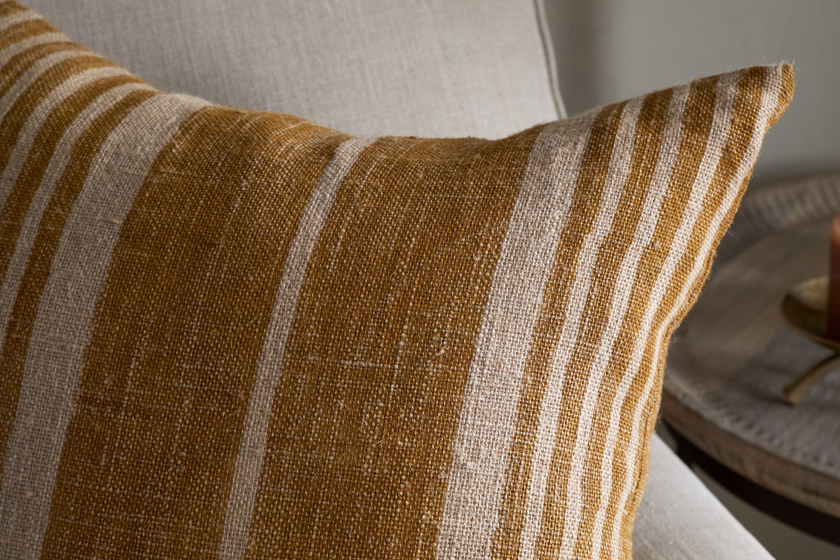 The Eesha Jute Cushion Cover - Ochre from nkuku rests on a light beige couch, its textured fabric with mustard yellow and beige stripes in focus. A round wooden side table with a candle appears in the background.