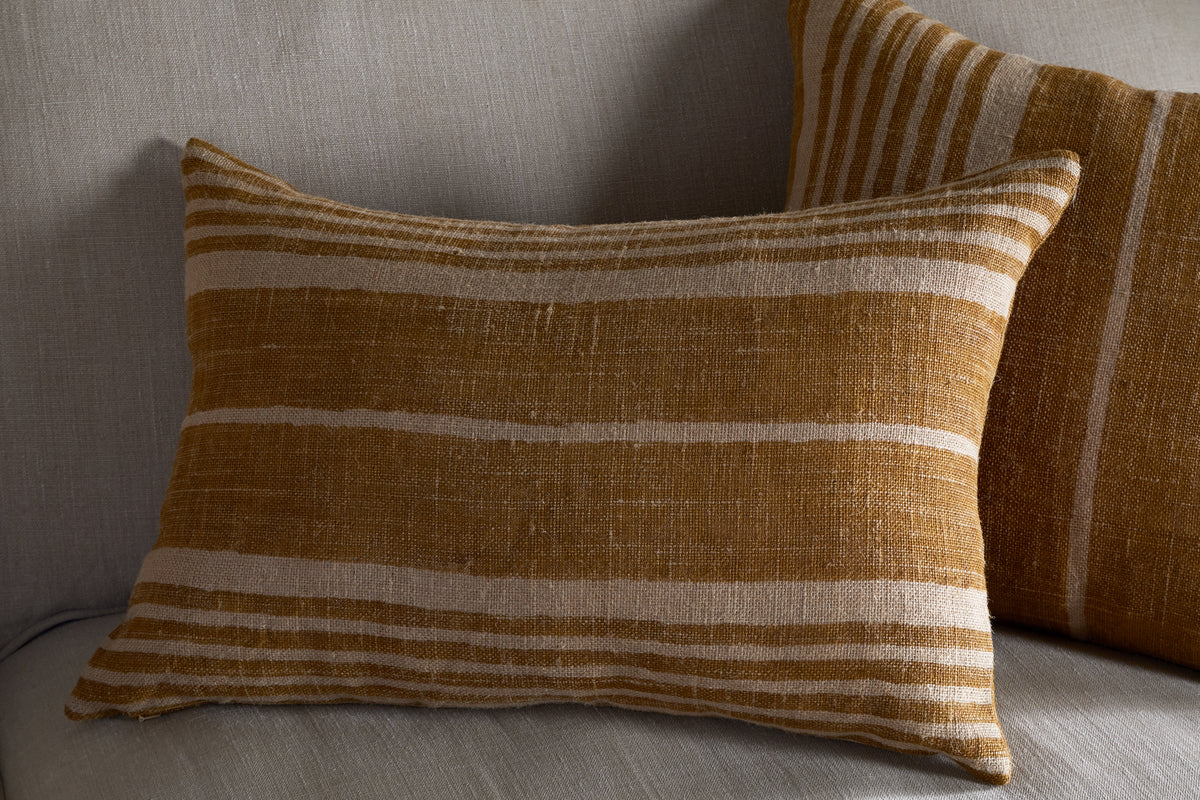 Two Eesha Jute Cushion Covers by nkuku in ochre with a modern rustic striped design rest on a light gray sofa, adding warm, cozy texture and color to the neutral background.