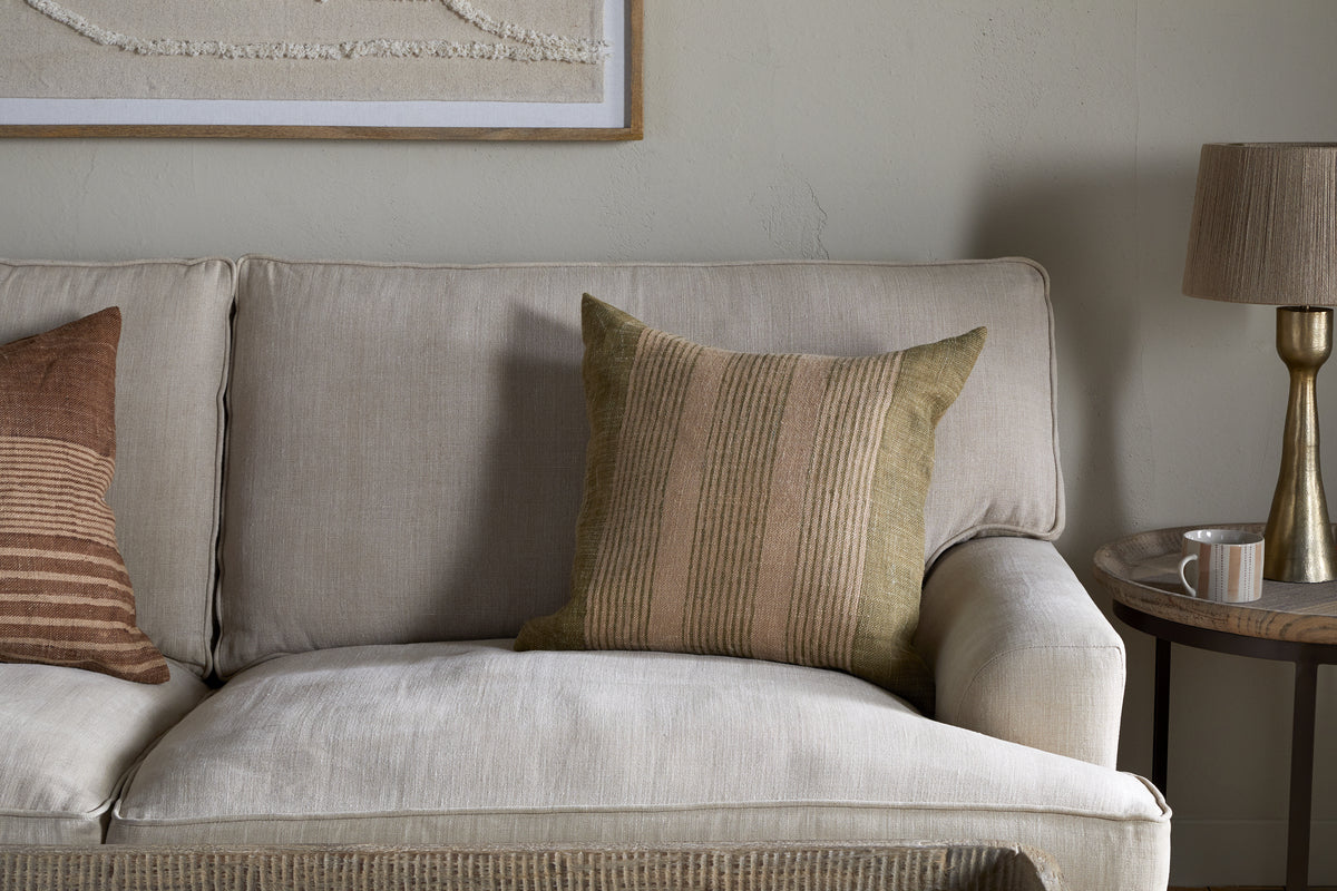 A beige sofa with the nkuku Eesha Jute Cushion Cover in sage green and a striped olive pillow sits by a round wooden side table holding a ceramic mug and brass lamp. Above, framed textured wall art completes the modern rustic décor.
