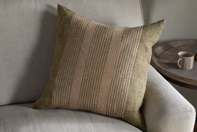 The nkuku Eesha Jute Cushion Cover in sage green rests against a light gray sofa, adding modern rustic charm. A round wooden side table with a beige mug nearby enhances the cozy, inviting atmosphere.