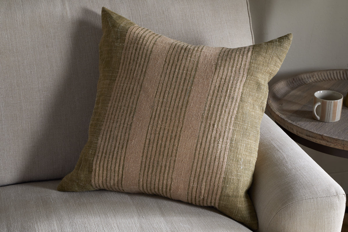 The nkuku Eesha Jute Cushion Cover in sage green rests against a light gray sofa, adding modern rustic charm. A round wooden side table with a beige mug nearby enhances the cozy, inviting atmosphere.