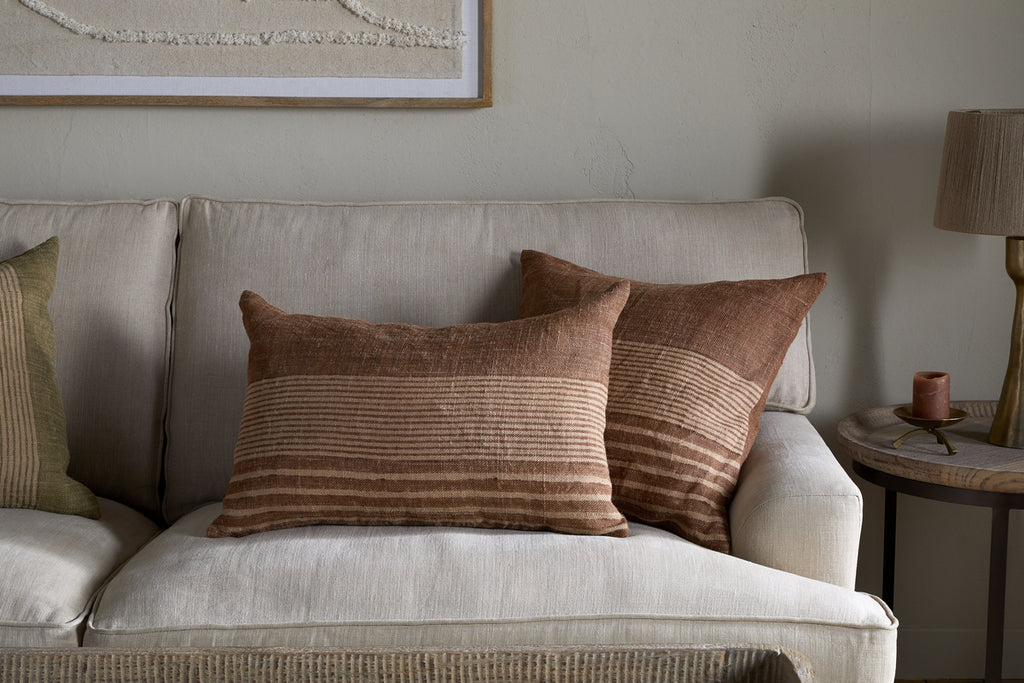 A cozy beige sofa is styled with modern rustic touches, including nkuku’s Eesha Jute Cushion Cover in Rust, brown striped and green pillows, a round wooden table with a lamp and candle, and textured wall art above in neutral tones.