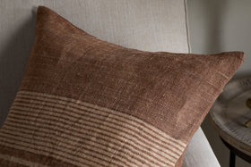 The Eesha Jute Cushion Cover - Rust by nkuku, featuring a brown textured finish with subtle beige stripes, rests on a light beige chair. Its modern rustic design adds warmth, complemented by a small round wooden side table in the background.