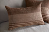 The Eesha Jute Cushion Cover - Rust by nkuku, with its textured, woven design and muted earthy tones, rests on a light gray fabric sofa. Partially visible behind it is another similar modern rustic cushion.