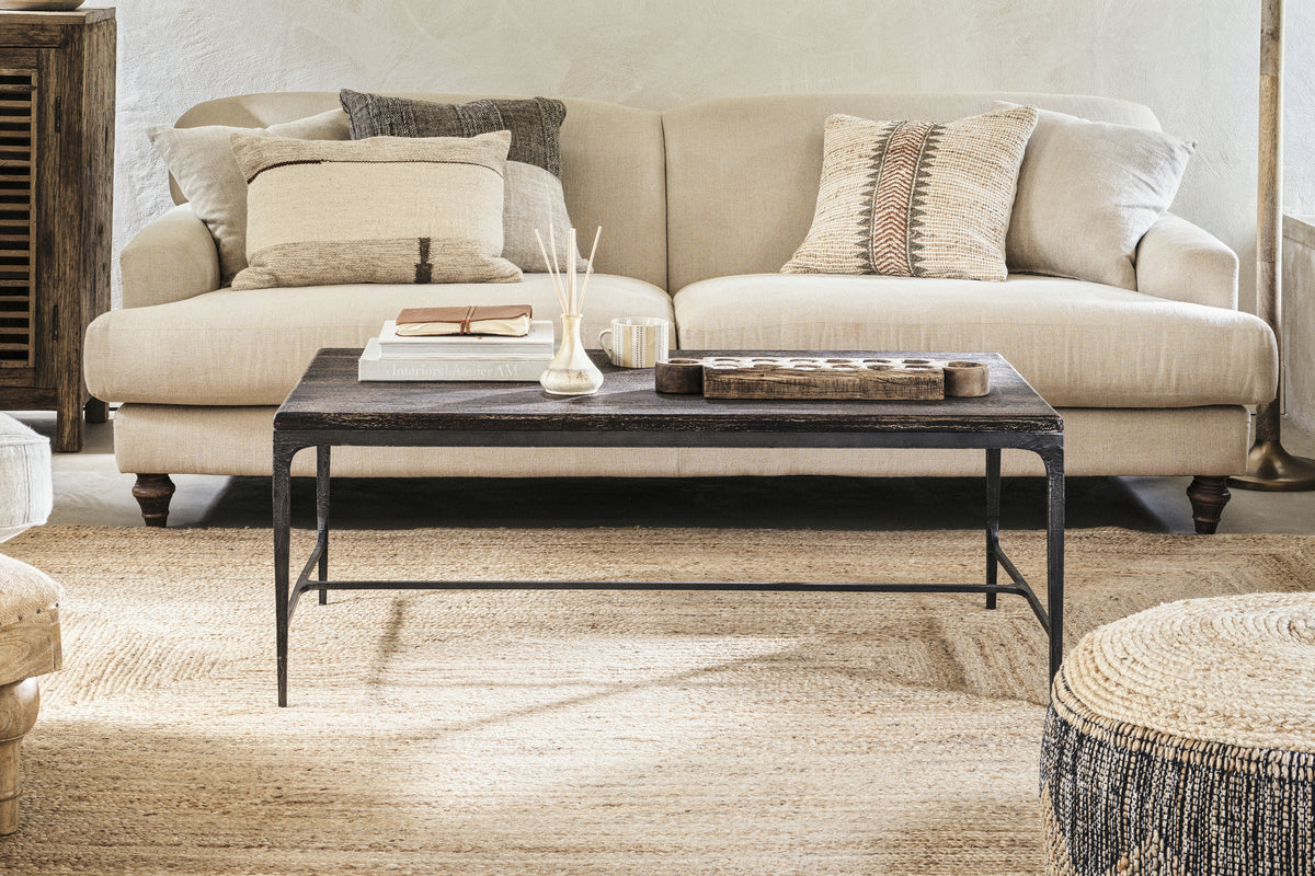 A beige sofa with patterned and plain cushions sits behind the nkuku Sita Reclaimed Wood & Iron Coffee Table in Chocolate Brown, which holds a tray, book, and reed diffuser. A jute rug and neutral decor add warmth to this cozy, modern living room.