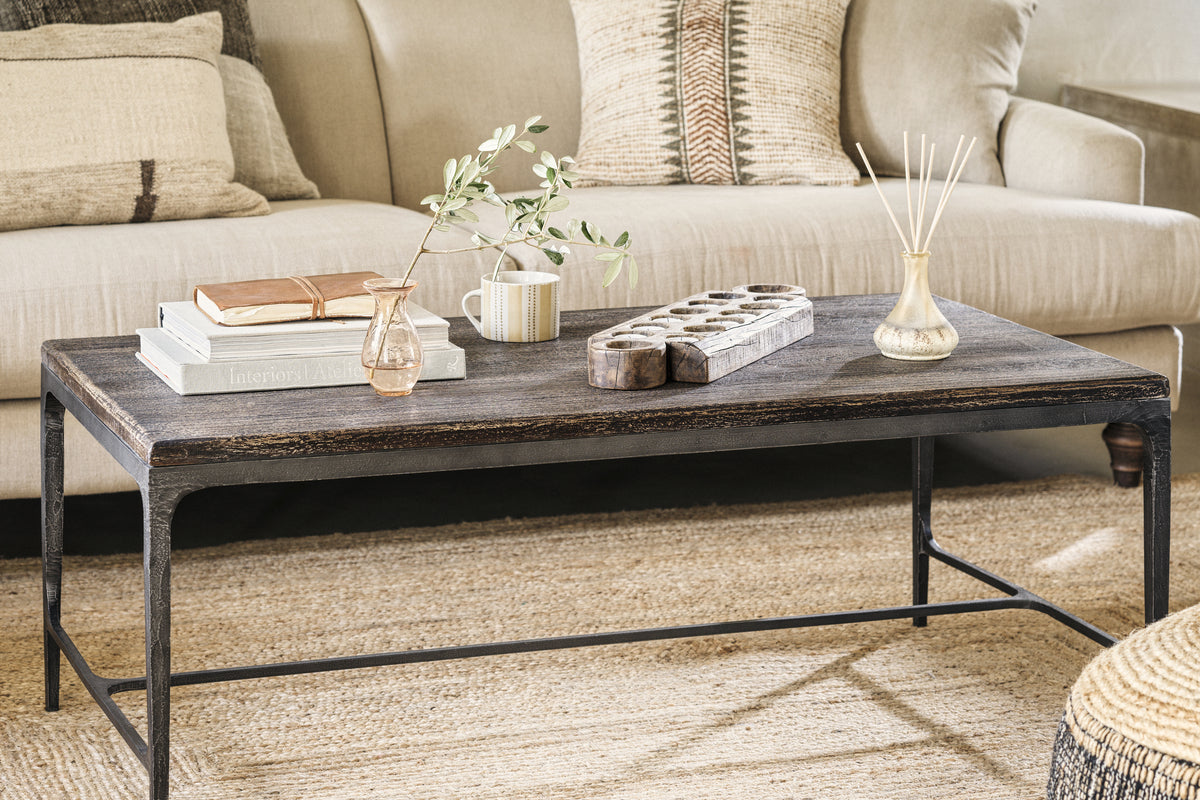 A cozy living room featuring the nkuku Sita Reclaimed Wood & Iron Coffee Table in Chocolate Brown, styled with books, a vase of greenery, a mug, wooden tray, and reed diffuser on a woven rug before a beige sofa with neutral-patterned pillows.