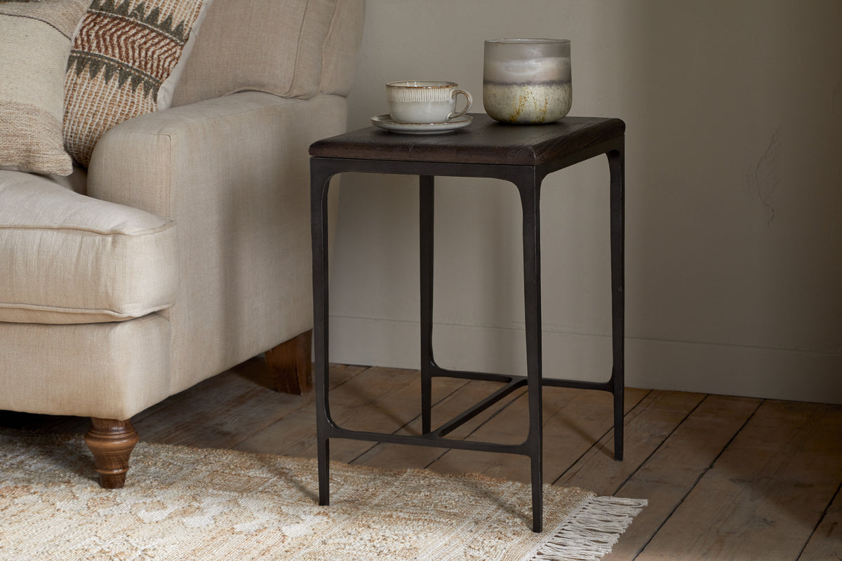 The nkuku Sita Reclaimed Wood & Iron Side Table in chocolate brown stands by a beige sofa topped with a ceramic teacup, saucer, and glass jar candle. A woven rug peeks out beneath, while textured cushions accent the sofa.