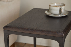 A close-up of the nkuku Sita Reclaimed Wood & Iron Side Table in chocolate brown features thin metal legs. A beige ceramic cup and saucer rest on top, with a beige sofa in the softly lit background—an inviting accent piece.