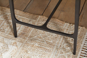 Close-up of the Sita Reclaimed Wood & Iron Side Table by nkuku, featuring a black iron leg resting on a textured beige woven rug. The rug overlays a chocolate brown reclaimed wood floor with visible grain and plank gaps.