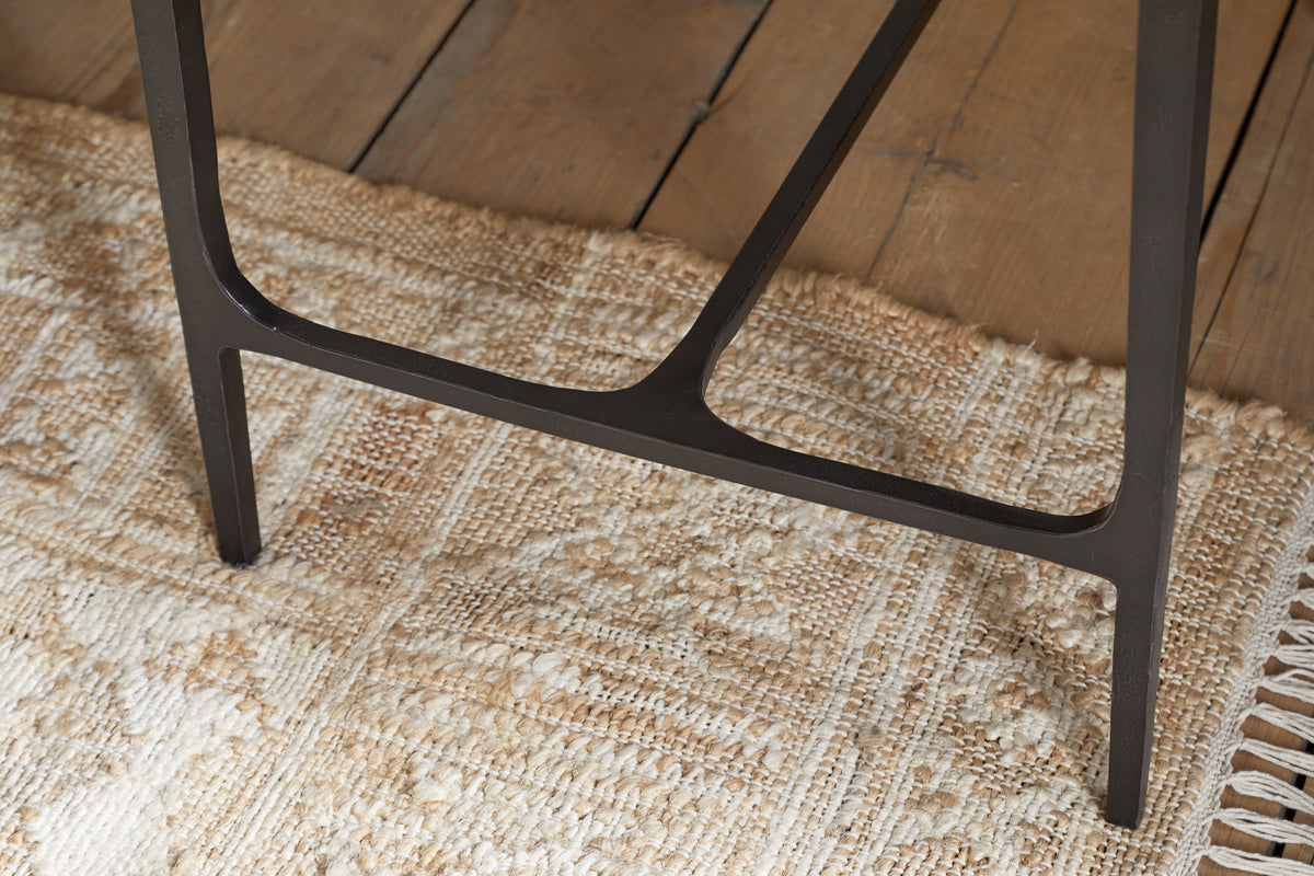 Close-up of the Sita Reclaimed Wood & Iron Side Table by nkuku, featuring a black iron leg resting on a textured beige woven rug. The rug overlays a chocolate brown reclaimed wood floor with visible grain and plank gaps.