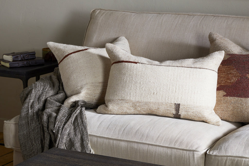 A cozy beige sofa features two nkuku Tamil Wool Cushion Covers in natural, with simple brown, charcoal, and rust tones. A gray woven throw is draped over the arm, and a side table with stacked books completes this warm, inviting setting.