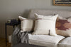 A beige sofa features three decorative pillows, including the nkuku Tamil Wool Cushion Cover in natural. A gray textured throw is draped over the arm. A small black side table holds stacked books beneath woven wall art.