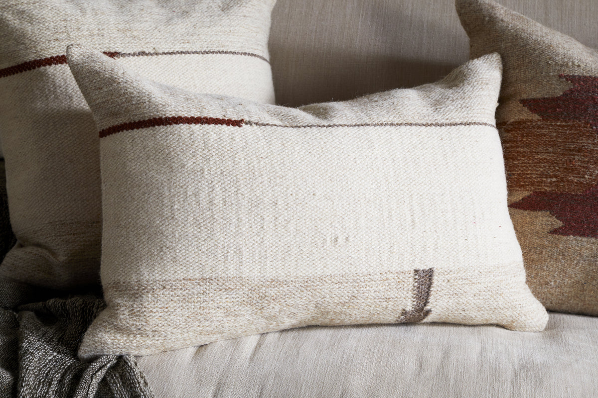The Tamil Wool Cushion Cover - Natural by nkuku features a cream, textured weave with subtle brown and tan stripes, adding minimal style to a beige sofa paired with charcoal and rust cushions and a gray throw blanket.