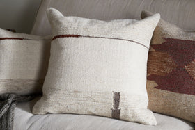 Three decorative cushions rest on a light beige sofa. The central cushion features the nkuku Tamil Wool Cushion Cover - Natural, with a minimal brown and dark red design, while charcoal and rust pillows add a cozy, rustic touch.