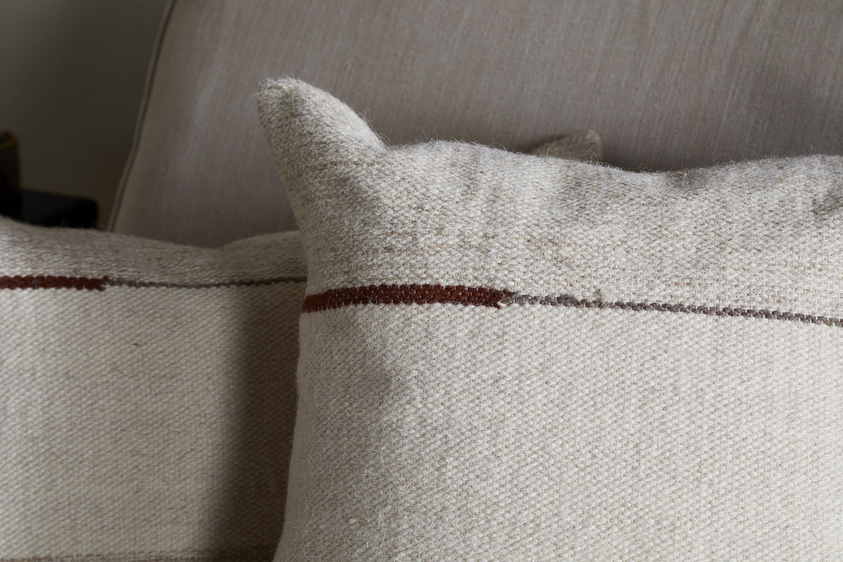 Two beige nkuku Tamil Wool Cushion Covers in natural, featuring a subtle woven texture and thin horizontal brown stripe, rest on a light sofa. Their minimal design adds a cozy, inviting touch to any space.