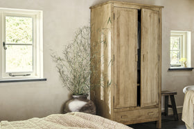 The nkuku Vivan Grooved Wardrobe - Natural with two doors stands by a big brown ceramic vase with tall branches. Cream walls, a beige knit blanket, black-framed windows, and a wooden stool complete the cozy bedroom storage setup.