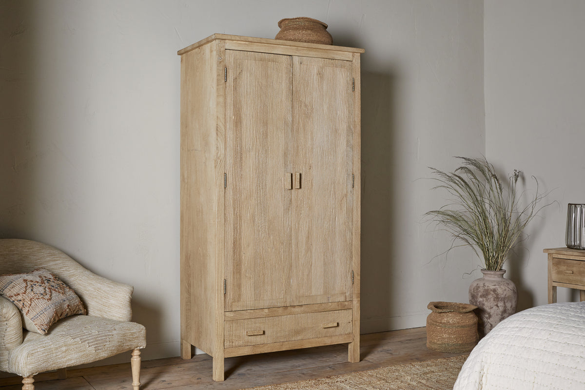 The nkuku Vivan Grooved Wardrobe - Natural, featuring two vertical doors and a lower drawer, stands in a neutral room with a woven basket on top. Nearby are a cushioned chair with pillow, a plant, another basket, and small table.