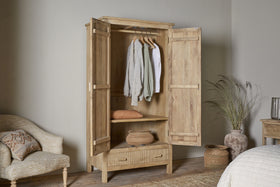 The nkuku Vivan Grooved Wardrobe - Natural features open doors displaying hanging clothes, a folded sweater, a woven basket on a shelf, and a closed drawer—offering elegant bedroom storage in serene neutral tones with soft natural light.