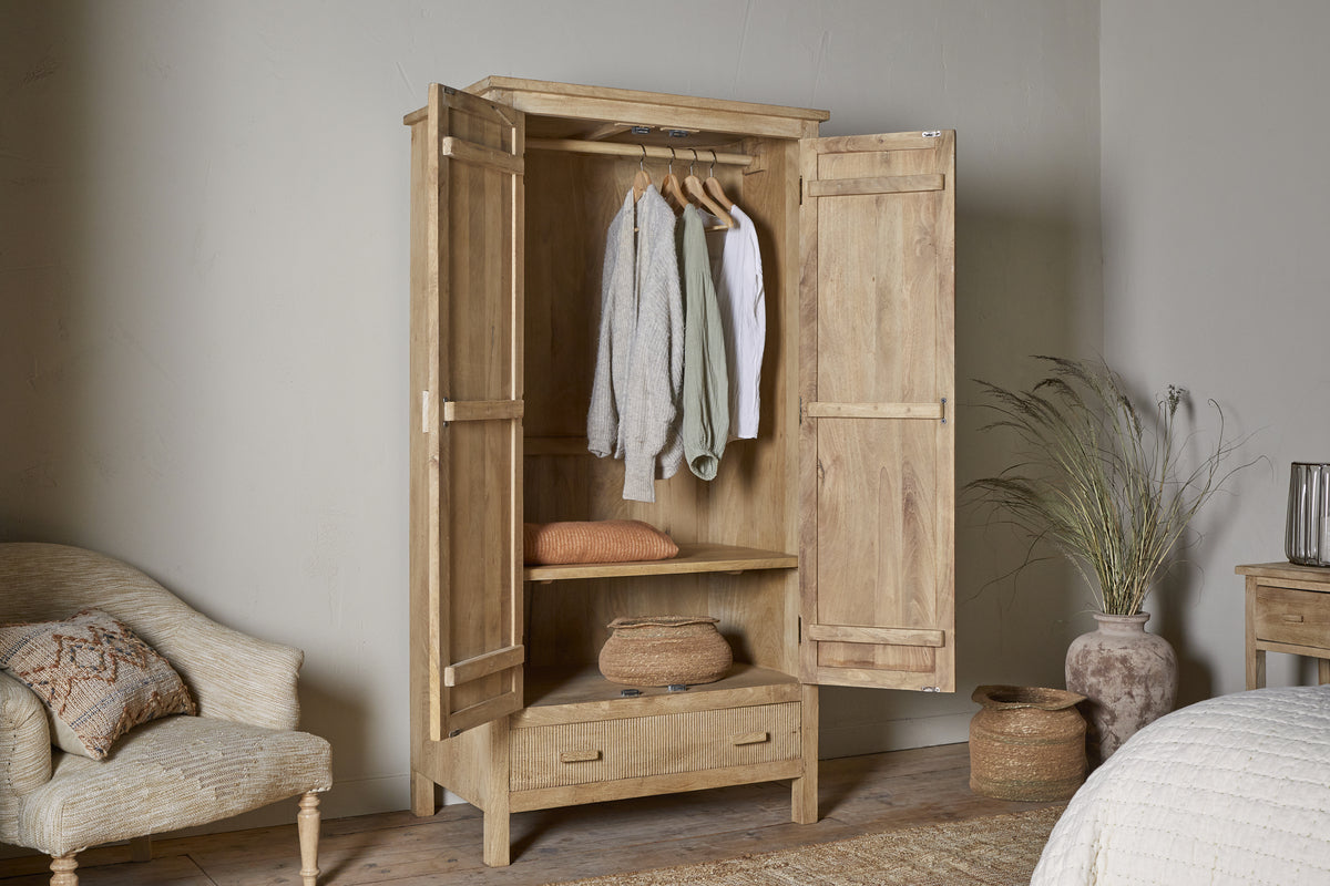 The nkuku Vivan Grooved Wardrobe - Natural features open doors displaying hanging clothes, a folded sweater, a woven basket on a shelf, and a closed drawer—offering elegant bedroom storage in serene neutral tones with soft natural light.
