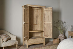 The Vivan Grooved Wardrobe - Natural by nkuku stands open in a softly lit bedroom, showing its two doors, interior shelf, and bottom drawer. Nearby are a cream armchair, woven basket with dried grass, and a light rug on wooden floors.