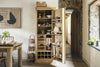 The nkuku Vivan Grooved Pantry Cupboard - Natural stands open in a rustic kitchen, showcasing jars and baskets on its shelves beside a wooden dining table with bread and plants near a stone archway, all bathed in natural light.