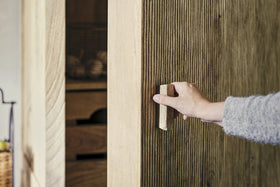 A person opens the nkuku Vivan Grooved Pantry Cupboard - Natural, featuring a textured ribbed wood surface and vertical handle. Inside the stylish wooden cabinet, shelves display baskets and storage boxes in the background.