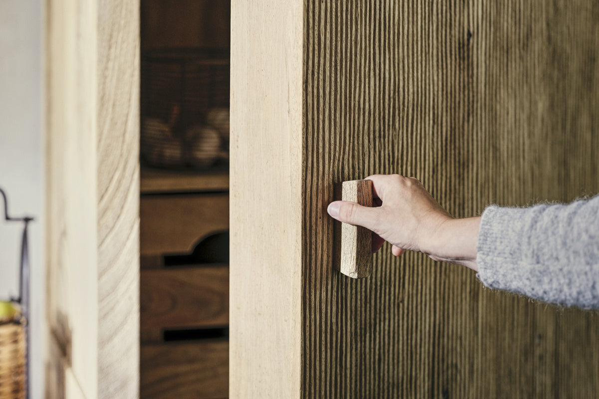 A person opens the nkuku Vivan Grooved Pantry Cupboard - Natural, featuring a textured ribbed wood surface and vertical handle. Inside the stylish wooden cabinet, shelves display baskets and storage boxes in the background.