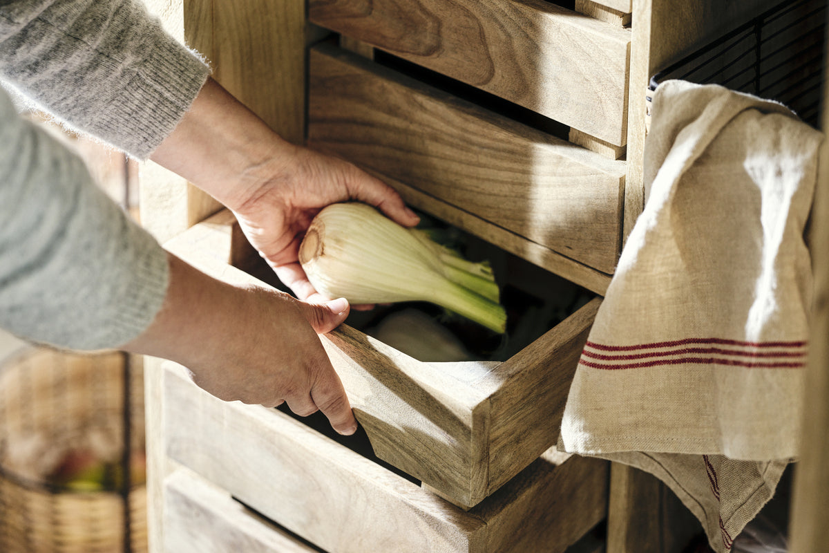 A person’s hands place a fresh fennel bulb into the Vivan Grooved Pantry Cupboard - Natural by nkuku, a rustic storage unit with multiple compartments. A beige kitchen towel with red stripes hangs at the side for country pantry charm.