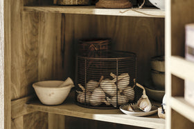 The nkuku Vivan Grooved Pantry Cupboard in natural wood brings charm to your kitchen, neatly storing a mortar and pestle, garlic bulbs in a wire basket, stacked ceramic bowls, and a plate of garlic cloves for a cozy, organized look.