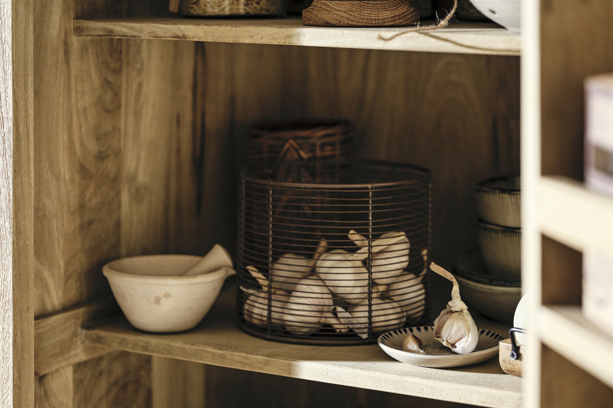The nkuku Vivan Grooved Pantry Cupboard in natural wood brings charm to your kitchen, neatly storing a mortar and pestle, garlic bulbs in a wire basket, stacked ceramic bowls, and a plate of garlic cloves for a cozy, organized look.