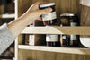 A hand in a gray sweater reaches to pick up a jar of orange marmalade from the Vivan Grooved Pantry Cupboard - Natural by nkuku, with other jars of preserves and boxes neatly arranged on the shelves in the background.